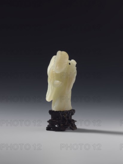 Monk with a rosary around his neck; unknown creator; ; figurine of a monk; Chinese; Qing dynasty 1644–1911; full sculpture; 18th century; material: sculpture; height 7.8 cm., A small carved pale yellow figure stands on a dark brown intricately carved base against a gradient background that shifts from dark gray at the top to a lighter gray near the bottom; the figure appears to have flowing robes and a rounded head with carved facial features, and a few subtle variations of creamy white and translucent yellow are visible in the material, while the base shows deep brown and almost black tones and casts a soft shadow on the gray surface.