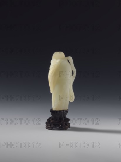 Monk with a rosary around his neck, unknown sculptor, figurine, monk subject, Chinese, Qing dynasty 1644–1911, full sculpture, 18th century, China, height 7.8 cm, A smooth, pale yellow carved object resembling a standing figure draped in flowing, rounded shapes rests on a dark, textured base that has an irregular, stacked appearance; the backdrop transitions from deep black at the top through dark gray to a lighter gray near the bottom, and a soft shadow extends from the base across the lighter gray surface.