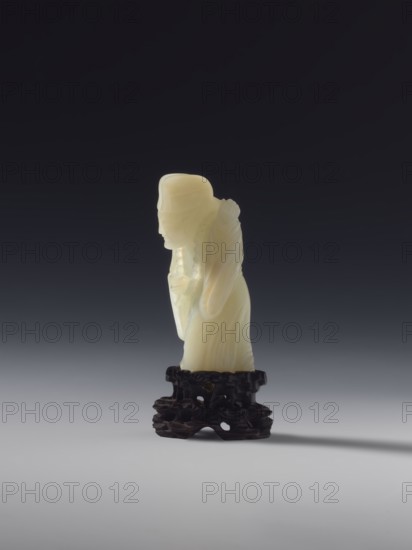 Monk with a rosary around his neck; unknown sculptor; figurine; Chinese; Qing dynasty 1644–1911; full sculpture; 18th century; medium: sculpture; height 7.8 cm., A pale cream-colored carved figurine of a person with a rounded head and draped clothing stands on a dark brown, intricately carved wooden base; the background is a smooth gradient from very dark near the top to lighter gray near the bottom, and there is a soft shadow cast to the right on the light gray surface.