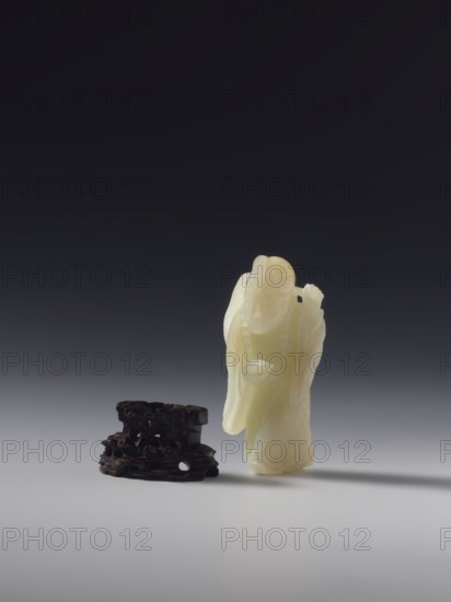 Monk with a rosary around his neck, unknown sculptor, figurine of a monk, Chinese, Qing dynasty 1644–1911, full sculpture, 18th century, China, height 7.8 cm, A pale cream-colored carved figure standing upright beside a dark brown, irregularly shaped base; the figure has flowing draped garments and a rounded head, with subtle glossy surface highlights and smooth contours, while the base has a rough, carved texture with hollows and a matte finish; the background transitions from very dark gray at the top to lighter gray near the bottom, and both objects cast soft shadows on the light gray surface beneath them.
