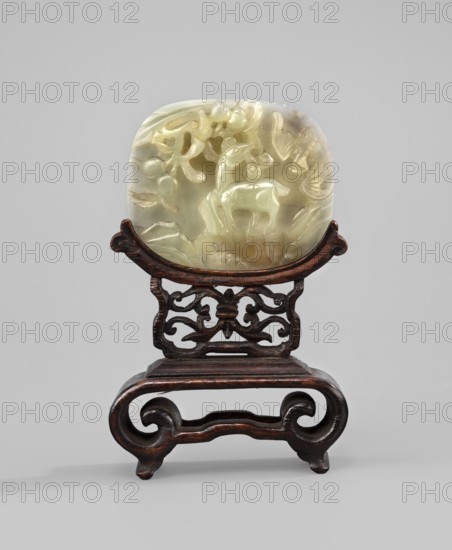 Small screen on base, Chinese art, Chinese culture, Qing 1644–1911, landscape motifs, peaches iconography, bats iconography, happiness symbol, deer; plaque of carved white jade; base of rosewood carved with an openwork pattern; 19th century, China; screen; height 11.8 cm., A carved pale green and creamy white translucent stone plaque with rounded edges sits on a dark brown carved wooden stand; the stone shows raised carved shapes including an animal figure and plant-like forms, with variations of pale yellowish-green and creamy white tones and some subtle brownish spots, while the wooden stand has a polished dark brown surface with carved scrollwork and curved legs.