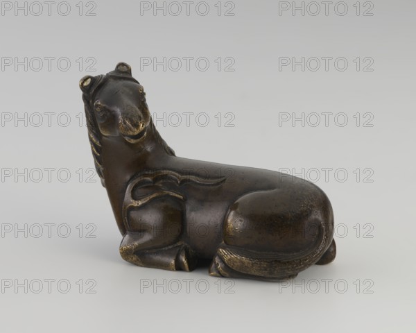 Paperweight — “heavenly horse”; creator, ; Chinese; Qing dynasty 1644–1911; 19th century; material/technique: sculpture/painting implements; subject: horse with flames iconography; form/function: figurine/paperweight; dimensions: height 5.7 cm, width 7.5 cm, depth 4.1 cm., A small sculpted animal figure shown in profile, rendered in dark brown and bronze tones with areas of worn golden highlights; the figure is reclining with a rounded body, a long neck, and a stylized head turned slightly upward, featuring carved ears and a snout; the surface includes carved decorative lines on the body and mane along the neck, and the legs are tucked under the torso with subtle textural wear on the feet and tail; the background is a smooth light gray that contrasts with the darker colors of the figure.