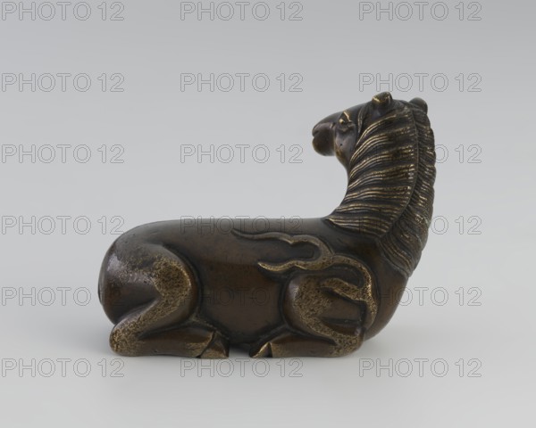 Paperweight — heavenly horse;, Chinese; Qing dynasty 1644–1911; 19th century; materials: painted sculpture painting implements; subject: horse with flames heavenly horse iconography; dimensions: height 5.7 cm, width 7.5 cm, depth 4.1 cm., A small sculpted animal figure shown in profile lying on a flat surface, with a rounded body, curled legs tucked beneath it, and an elongated neck turned slightly upward; the surface appears metallic with a dark brown patina and highlighted areas of golden bronze, the mane is textured with parallel grooves creating a ribbed pattern, and the figure shows smoother worn areas and subtle surface variations in shades of brown and gold against a neutral light gray background.