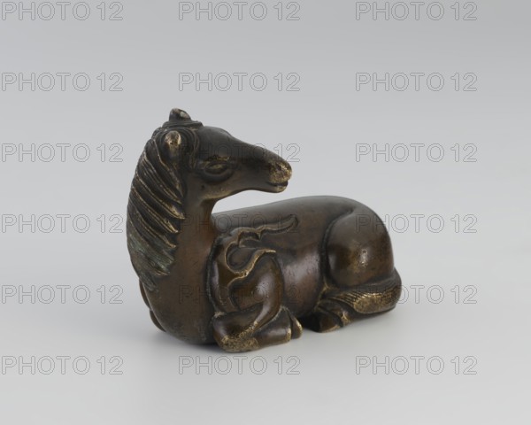 Paperweight — heavenly horse; maker, ; Chinese art; Chinese; Qing dynasty 1644–1911; 19th century; materials/objects: figurine sculpture functioning as a paperweight; subject: horse with flames heavenly horse motif; related to painting implements; dimensions: height 5.7 cm, width 7.5 cm, depth 4.1 cm., A small sculpted animal figure shown in a reclining pose with a turned head, rendered in dark brown and bronze tones with highlights of gold and hints of greenish patina; the surface shows smooth rounded forms and carved ridges suggesting a mane and stylized body details, set against a pale gray background.