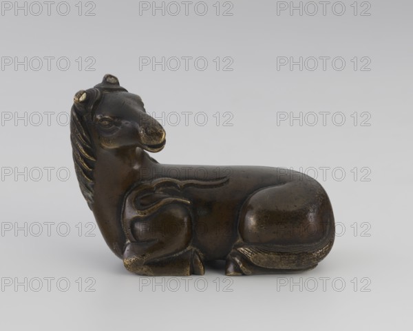 Paperweight — heavenly horse;, ; Chinese art; Chinese culture; Qing dynasty 1644–1911; 19th century; figurine paperweight depicting a horse with flame motifs; painted sculpture; materials include painting implements; dimensions: height 5.7 cm, width 7.5 cm, depth 4.1 cm., A small sculpted animal figure shown in profile, reclining with its legs tucked under its body and its head turned slightly upward; the surface appears metallic with dark brown and bronze tones with areas of lighter golden wear; the mane is stylized with carved ridges, and there are rounded ears and a pronounced snout; the body is smooth with subtle carved lines indicating limbs and a curled tail resting against the haunch; the object sits on a neutral light gray background.
