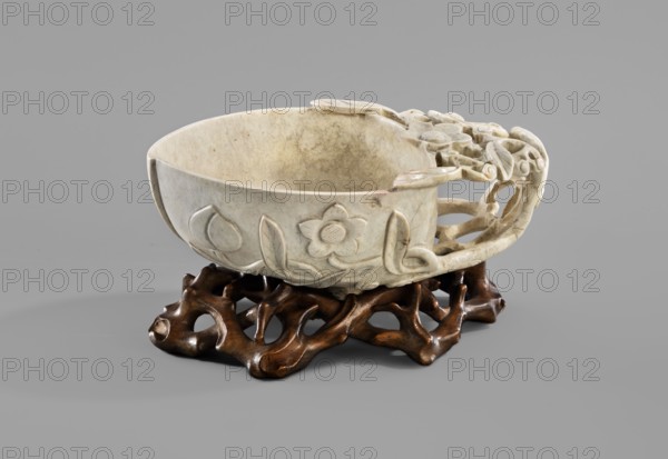 Bowl in the shape of half a peach on a stand., Chinese, Qing dynasty 1644–1911. 17th–18th century. Stone bowl with an openwork handle in the shape of a leafy flower branch. Wooden openwork stand with motif of woven branches and a five-petaled flower in the center. Subject/iconography: peaches. Type: vessel, bowl. Dimensions: bowl height 5 cm, width 13.5 cm; stand height 2.5 cm, width 12.3 cm., A carved pale stone bowl with floral and leaf motifs rests on an organic dark brown wooden stand; the bowl has an openwork carved handle with rounded, curved shapes and a smooth interior showing natural veining, and the scene sits against a uniform gray background with soft shadows.