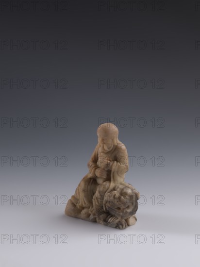 Luohan on a lion, arhat iconography, lion iconography, figurine, Chinese art, Qing 1644–1911, Chinese Buddhism, 19th century, China, sculpture; height 9.3 cm, width 7.7 cm, depth 3.7 cm, A small carved figure made of mottled beige and light brown stone sits on a smooth surface, showing a seated person with draped robes and a rounded, smooth head; the figure's hands are positioned near the torso and the robes have flowing folds. The person sits atop or beside a carved animal-like form with a rounded face and visible carved features such as a snout and paws. The stone shows natural speckling and subtle variations in beige, cream, tan, and darker brown tones, and the background is a smooth gradient ranging from dark gray at the top to pale gray near the bottom.