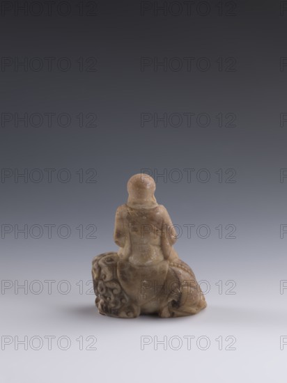 Luohan on a lion;, arhat iconography; lion iconography; figurine; Chinese art; Chinese culture; Qing dynasty 1644–1911; Chinese Buddhism; 19th century; China; sculpture; height 9.3 cm; width 7.7 cm; depth 3.7 cm., A small carved figurine in beige and light brown tones sits on a pale surface against a gradient gray background, showing the back view of a seated figure with rounded head and draped clothing; the base includes carved, swirling decorative forms and textured areas with subtle darker brown speckles and patches.