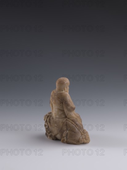 Luohan on a lion;, arhat iconography; lion iconography; figurine; Chinese art; Qing dynasty 1644–1911, 19th century, China; Chinese Buddhism; sculpture; height 9.3 cm, width 7.7 cm, depth 3.7 cm., A small carved figure in a warm tan and brown stone sits on a smooth surface, shown from the back; the figure appears robed with folds and rounded shoulders, a rounded head, and a textured base that includes carved ornamental details and a beaded element, with subtle lighter speckles and variations in the stone; the background is a smooth gradient of dark gray at the top fading to lighter gray toward the surface, and the surface beneath the figure is a pale gray.