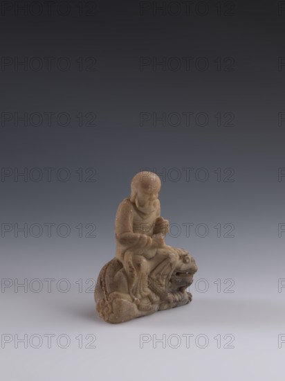 Luohan on a lion, arhat iconography, lion iconography, figurine, Chinese art, Chinese culture, Qing dynasty 1644–1911, Chinese Buddhism, 19th century, China, sculpture; height 9.3 cm, width 7.7 cm, depth 3.7 cm., A small carved figurine in shades of tan and light brown sits on a smooth pale gray surface against a gradient background that shifts from dark gray at the top to medium gray near the center; the figurine depicts a seated human figure with a rounded head and draped clothing, hands positioned in front, seated atop an animal-like form with a pronounced open mouth and rounded features; the carving shows smooth, worn textures and subtle speckled markings across its surface, with shadows beneath and to one side.