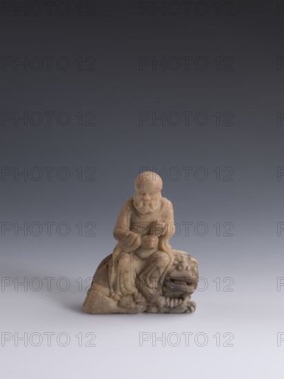Luohan on a lion;, arhat and lion iconography; figurine; Chinese art; Qing dynasty 1644–1911, 19th century; China; Chinese Buddhism; sculpture; height 9.3 cm, width 7.7 cm, depth 3.7 cm., A small carved figure in shades of beige and light brown sits on a surface against a smooth gradient background that transitions from dark gray at the top to lighter gray near the bottom; the figure depicts an elderly person with a rounded head and a pronounced beard, wearing flowing robes with carved folds and holding an object in their hands, seated atop a stylized creature with a broad face, open mouth, visible teeth, and rounded features; the carving shows subtle surface texture and small speckles of darker brown throughout, and the overall tones include beige, light brown, dark gray, and lighter gray.