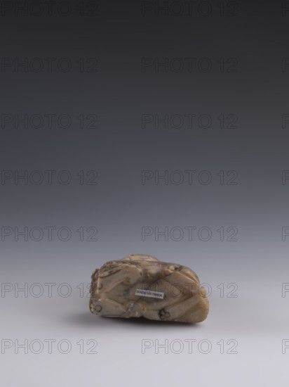 Luohan on a lion;, arhat iconography; lion iconography; figurine; Chinese art; Chinese culture; Qing dynasty 1644–1911; Chinese Buddhism; 19th century; China; sculpture; height 9.3 cm, width 7.7 cm, depth 3.7 cm., A small carved object resting on a smooth surface, showing a rounded shape with carved indents and raised areas; the object appears in shades of beige, tan, and light brown with some darker brown veins and spots, and it casts a soft shadow beneath it; the background transitions from light gray near the surface to dark gray higher up.
