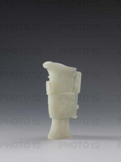 Vase with two taotie masks. Chinese, Qing dynasty 1644–1911, 18th–19th century. Porcelain vessel with full sculpture and bas-relief decoration depicting taotie masks and dragons. Height 15.2 cm; width 8.8 cm; depth 3.3 cm., A pale, smooth carved object with a glossy, translucent surface stands upright against a gradient background. The object has an elongated base that flares slightly and tapers into a wider midsection decorated with subtle carved patterns, then narrows and rises into a curved, spouted top with a rounded lip. Small protruding tabs and pierced sections appear on the sides of the midsection, and faint incised lines and motifs decorate various surfaces. The predominant color of the object is a milky off-white, and the background shows gradations of dark gray at the top blending into lighter gray toward the bottom.