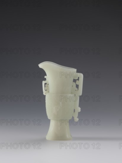 Vase with two taotie masks; Chinese porcelain vessel, Qing dynasty 18th–19th century; decoration: dragons and taotie masks in full sculptural and bas-relief; dimensions: height 15.2 cm, width 8.8 cm, depth 3.3 cm; origin: China; cultural attribution: Chinese; function: vase., A pale, translucent vessel in a white to very light green hue sits centered on a smooth surface against a dark gray to gradient background, with a curved spout at the top and a flared pedestal base; the body of the vessel shows carved decorative motifs and stylized handles on either side with pierced elements, and the overall surface has a smooth, slightly glossy finish.