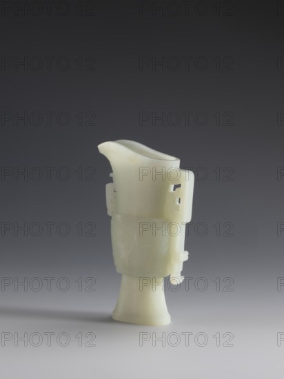 Vase with two taotie masks;, Chinese, Qing dynasty 1644–1911; 18th–19th century; porcelain vessel with dragons and taotie iconography in full sculpture and bas-relief; height 15.2 cm, width 8.8 cm, depth 3.3 cm., A pale, creamy white carved object resembling a slender vessel stands upright against a smooth gradient background that shifts from dark gray at the top to lighter gray near the bottom; the vessel has a flared base, a slightly curved body with subtle carved decorative patterns, and a fluted upper section that narrows into a curved spout, with small rectangular openwork handles on its sides creating a faint shadow on the surface beneath.