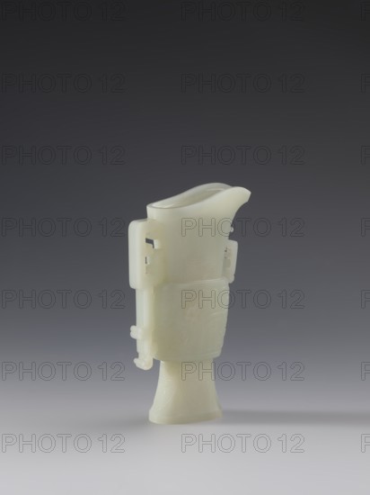 Vase with two taotie masks; Chinese; Qing dynasty 1644–1911; 18th–19th century. Porcelain vessel with full sculpture and bas-relief decoration featuring dragons and taotie mask iconography. Height 15.2 cm; width 8.8 cm; depth 3.3 cm., A pale off-white carved object stands upright against a smooth gradient background that shifts from dark gray at the top to light gray at the bottom. The object has a flared base tapering into a cylindrical midsection with a raised rectangular band and a slightly flared, boat-shaped top with a curved pouring lip. A few carved projections and loop-like handles extend from the sides, and subtle incised patterns and shallow relief decorations cover parts of the surface. The object casts a soft shadow on the lighter foreground. The visible colors are off-white, dark gray, and light gray.