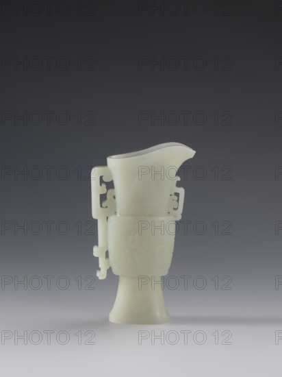 Vase with two taotie masks; maker,  from Ignacy Jan Paderewski; Chinese, Qing dynasty 1644–1911; 18th–19th century; porcelain vessel with dragons and taotie iconography in full sculpture and bas-relief; height 15.2 cm, width 8.8 cm, depth 3.3 cm., A pale, smooth vessel in a white to creamy color sits centered against a gradient background of dark gray at the top fading to lighter gray toward the bottom. The vessel has a flared mouth with a curved pouring spout, an elongated body with carved decorative patterns, and an attached ornate handle with angular cutouts and a hooked end. The base narrows then flares outward, and subtle carved motifs wrap around the midsection and upper portion of the piece. The surface appears polished and translucent in places, casting soft shadows on the light gray surface beneath it.