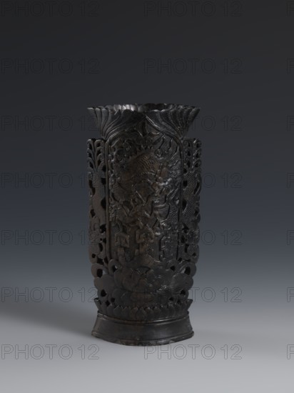 Vase, gift; Chinese, Qing dynasty 1644–1911; dark soapstone vase with bas-relief and openwork in places, decorated with dragons; one of three; 19th century; vessel; height 17.7 cm, width 8.7 cm., A tall, dark carved object with a flared top and a rounded base stands against a gradient gray background; the surface shows intricate openwork and relief carving with curving, floral and organic motifs, including petal-like shapes near the base and layered, leaf-like forms toward the rim, and the color palette is primarily dark brown to nearly black with subtle variations in sheen.