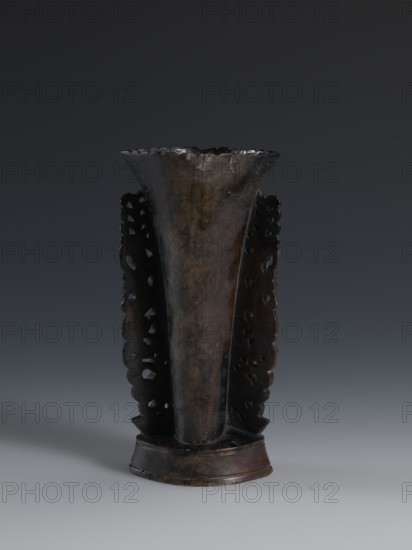 Vase, gift, Qing dynasty 1644–1911, 19th century, China; dark soapstone vase with bas-relief and openwork in places, featuring dragons iconography; one of three; height 17.7 cm, width 8.7 cm., A tall, vase-like object with a flared, scalloped rim and a narrow base stands against a dark gray background; the object has a vertical, slightly tapered central body flanked by two decorative, pierced panels with scalloped edges and irregular cutouts, and the surface shows mottled shades of dark brown, black, and hints of olive with a subtle glossy sheen.