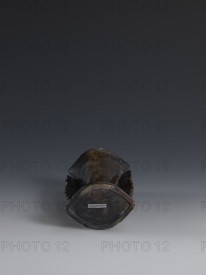 Vase, Qing dynasty 19th century, China. Dark soapstone vase with bas-relief and openwork in places, decorated with dragons. One of three. Height 17.7 cm; width 8.7 cm., A dark, elongated object lies centered on a smooth surface, positioned so its rounded end faces the viewer; the object has a worn, textured surface with tones of dark brown, black, and hints of muted bronze, and there are darker shadowed areas along its sides where carved or ridged details create a scalloped outline; the background transitions from a deep charcoal at the top to a lighter gray near the bottom, and a soft shadow falls to the left side of the object on the surface.