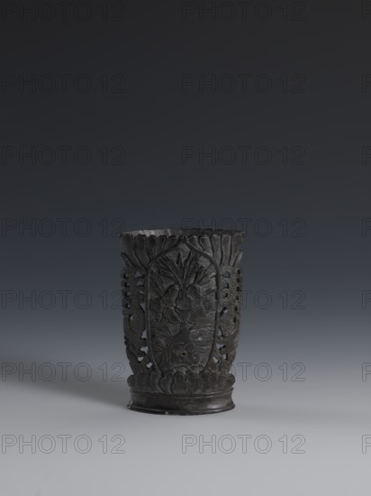 Vase, gift, vase, Chinese art, Chinese culture, Qing 1644–1911, plant ornament, vase made of dark soapstone, bas-relief, openwork in places; one of three, 19th century, China, vessel, height 11.3 cm, width 7.8 cm, A dark carved cylindrical object sits centered against a gradient background, showing a few shades of gray and black in the surroundings; the object itself is mainly black with subtle dark gray highlights and a slightly lighter gray base. The surface of the object displays carved floral and leaf patterns with openwork sections and scalloped edges along the top and bottom. The background transitions from very dark gray at the top to lighter gray near the bottom, creating a muted contrast with the black carved piece.