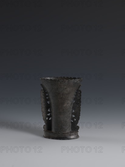Vase, made of dark soapstone with bas-relief and openwork in places; plant ornament. One of three. 19th century, Qing 1644–1911, China. Vessel. Height 11.3 cm; width 7.8 cm., A dark, weathered vase-like object with a flared rim and scalloped edge stands centered on a smooth, light-gray surface against a gradual dark-gray to nearly black background; the object has a vertical, slightly tapered body with a textured, worn surface in shades of dark gray and black, and features a pair of ornate, pierced side panels with irregular cutouts and scalloped outlines, casting a faint shadow to the left on the surface.