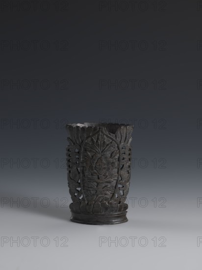 Vase, flattened vase made of dark soapstone with bas-relief and openwork in places, engraved. One of three. 19th century, China. Vessel; height 11.2 cm, width 7.8 cm, depth 3.3 cm. Qing 1644–1911. Chinese, plant ornament., A dark carved vessel sits centered against a muted gradient background, showing a rounded base and a slightly flared rim with scalloped edges; the vessel's surface features intricate raised floral and foliate patterns with openwork sections on the sides, and the overall visible color tones are dark gray to black with subtle lighter gray highlights where light hits the carved details.