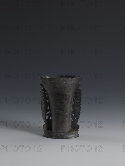 Vase, flattened vase made of dark soapstone with bas-relief and openwork in places, engraved. One of three. 19th century, China. Qing 1644–1911. Plant ornament. Vessel, height 11.2 cm, width 7.8 cm, depth 3.3 cm., A dark, weathered metal vessel with a flared, scalloped rim sits centered on a smooth surface against a gradient gray background; the vessel has a tapered body that widens at the top, decorative pierced side panels with ornate cutouts, and a slightly raised circular base, showing a patina with tones of dark gray, charcoal, and subtle brown.