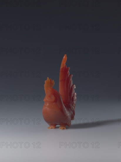 Rooster;, sculptor; ; Chinese art; Chinese culture; Qing dynasty 1644–1911; figurine of a rooster; 18th–19th century; China; sculpture; height 8.5 cm; width 7.0 cm; depth 2.6 cm, A small sculpted rooster figure in shades of red and orange stands on a smooth surface, facing slightly left, with a tall, detailed tail rising behind it; the background is a gradient of dark gray to lighter gray near the bottom, and a soft shadow extends to the right of the figure.