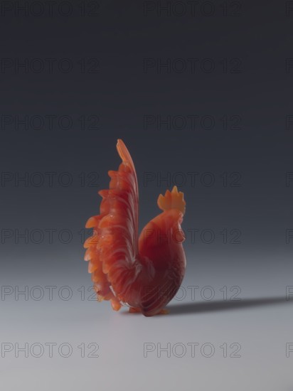 Rooster; maker, sculptor; gift; Chinese art; Chinese culture; Qing dynasty 1644–1911; figurine of a rooster iconography; 18th–19th century, China; sculpture; height 8.5 cm, width 7.0 cm, depth 2.6 cm, A glossy carved rooster figurine in shades of red and orange stands on a smooth light-gray surface against a dark gray to near-black gradient background, showing a tall fanned tail with layered feathers and a crest on its head, with soft highlights and a subtle shadow extending behind it.