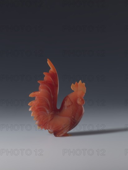 Rooster; sculptor, Chinese; Qing dynasty 1644–1911; 18th–19th century; figurine of a rooster; sculpture; height 8.5 cm, width 7.0 cm, depth 2.6 cm., A carved rooster figurine made of a smooth, translucent material in shades of red and orange stands on a flat surface, with a curved tail fanning upward and detailed feather texture along its body; the background transitions from dark gray at the top to lighter gray near the bottom, and a soft shadow extends beside the figurine.