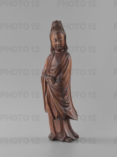 Guanyin Bodhisattva, unknown sculptor; figurine; Chinese art; Chinese Buddhism; bodhisattva iconography; goddess; Qing dynasty 1644–1911; 19th century; wood carving, solid, polished; height 32 cm; sculpture., A carved wooden standing figure with a smooth brown finish wearing flowing draped robes, a headdress, and a serene facial expression; the figure's hands are positioned near the waist, the robes show carved folds and drapery, and the feet are visible at the base. The background is a uniform light gray. Colors visible include shades of brown on the figure and gray in the background.