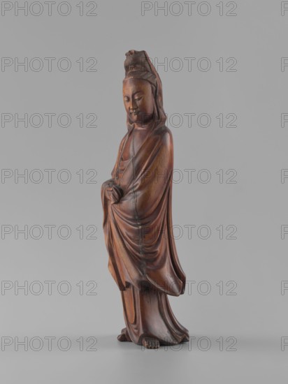 Guanyin Bodhisattva, unknown sculptor, figurine, Chinese, Qing dynasty 1644–1911, 19th century, wood carving, solid polished, bodhisattva iconography, goddess, Chinese Buddhism, height 32 cm., A carved wooden figure of a robed person standing on a flat surface, shown against a plain gray background; the figure has a smooth brown finish with visible wood grain and darker brown shading in folds and creases, a serene facial expression with closed eyes, a headdress, draped garments with flowing folds, hands held together near the waist, and bare toes visible at the base.