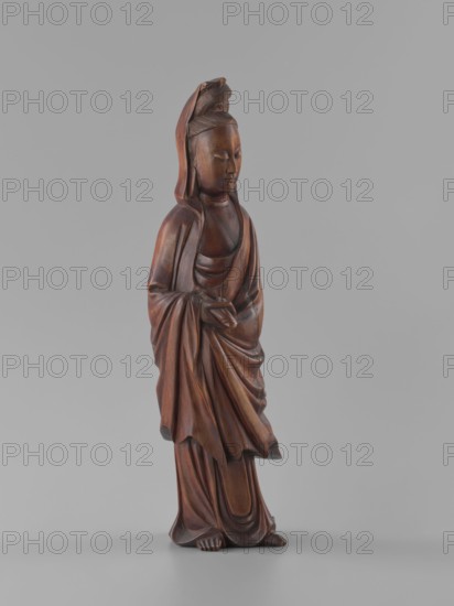 Guanyin Bodhisattva, unknown sculptor, gift, figurine, Chinese art, Chinese culture, goddess, Chinese Buddhism, Qing 1644–1911, bodhisattva iconography, wood carving, solid, polished, 19th century, China, sculpture, height 32 cm, A carved wooden figure of a robed person standing on a flat surface, depicted with closed eyes and a serene facial expression, wearing a hood or head covering with a carved detail at the top, draped garments with flowing folds and layered robes, hands held together at the waist, exposed feet peeking from beneath the robes; the wood shows warm brown tones with darker and lighter brown variations and subtle reddish-brown highlights against a plain neutral gray background.