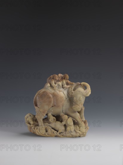 Buffalo with calves and shepherds, sculptor unknown. Sculpture of decorative Fushan Mountain Zhejiang stone soapstone with natural patina. Chinese; Qing dynasty 1644–1911, 18th–19th century. Subject: buffaloes with calves and shepherds. Height: 16.5 cm., A carved stone sculpture depicting a buffalo with curved horns standing on a textured base, with a group of small human figures riding on its back and clustered around its legs; the sculpture shows smooth and detailed carving with rounded forms and flowing shapes, and the stone displays a range of beige, tan, brown, and cream tones with subtle variations and darker patches, set against a gradient background that transitions from dark gray at the top to lighter gray toward the bottom.