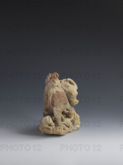 Buffalo with calves and shepherds, unknown creator sculptor, buffaloes, Chinese art, figurines, Chinese culture, Qing 1644–1911, sculpture made of decorative Fushan Mountains Zhejiang stone soapstone, full, natural patina, 18th–19th century, China, sculpture, height 16.5 cm, A small carved sculpture of an animal resembling a bull with curled horns stands on an irregular base, showing a rounded body, muscular legs, and a lowered head; several smaller animal figures cluster around its feet and along the base, merging into flowing carved forms. The surface shows a mix of warm beige, pale yellow, light brown, and darker brown tones with subtle mottling and smooth polished areas. The sculpture is positioned on a smooth, light gray surface against a dark gray to nearly black gradient background.