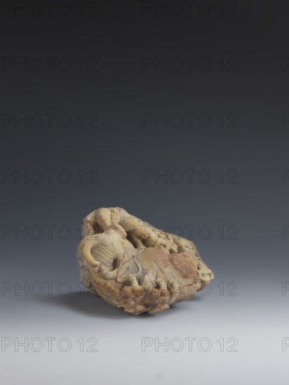 Buffalo with calves and shepherds, sculpted from decorative Fushan Mountains Zhejiang stone soapstone, natural patina; figurine in Chinese Qing dynasty style 1644–1911, 18th–19th century; sculpture, height 16.5 cm., A carved stone object sits on a smooth surface against a dark to light gray gradient background; the object is irregularly shaped with intricate relief carving showing curved and rounded forms, organic folds, and coiled shapes, and it displays a range of colors including beige, cream, light brown, warm tan, and hints of pale yellow, with some darker brown and reddish-brown areas and subtle shadows beneath it.