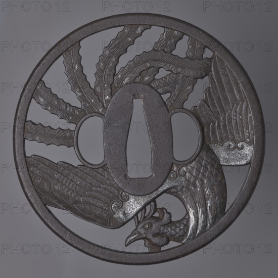 Crossguard tsuba, Kinai,  school, Echizen Kinai,  school; phoenixes hoo/hôô iconography; sword crossguard for katana, melee weapon; Japanese, Edo period ca. 1800, 1603–1868; Tsuba round maru gata with a rectangular edge with slight convexity kaku mimi ko-niku; oval seppa-dai; oval ryôhitsu-ana matching the usual kozuka-hitsu-ana; decoration: mythical bird hôô with spread wings and long tail feathers arranged in a fan-like pattern incorporated into a circle on both sides; plumage details engraved; diameter 9.3 cm; thickness 0.5 cm., A round metal disk with a dark brown and gray patina featuring an openwork design of a bird with outstretched, textured wings and a detailed head near the lower edge, surrounded by a circular rim and a central oval opening flanked by two smaller openings, with carved feather patterns and subtle scratches and lighter metallic highlights on raised areas.