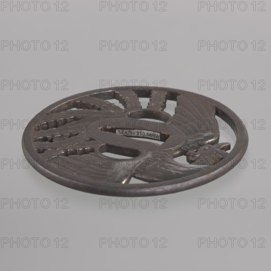 Crossguard tsuba, Kinai,  school author, Echizen Kinai,  school; phoenixes iconography, hoo phoenix; sword crossguard for katana; Japanese art, Japanese culture; weapon, melee weapon; Edo 1603–1868 swords; Tsuba round maru-gata with a rectangular edge with slight convexity kaku mimi ko-niku; oval seppa-dai; oval ryôhitsu-ana matching the usual kozuka-hitsu-ana; decoration with a mythical bird hôô with spread wings and long tail feathers arranged in a fan-like pattern, incorporated into a circle on both sides; feather details engraved. Ca. 1800 Edo Period, 1603–1868, Japan. Tsuba; diameter 9.3 cm; thickness 0.5 cm., A round metal object with a dark brown and black patina sits against a light gray background, featuring carved openwork sections and a raised outer rim; the surface shows textured feather-like patterns and scalloped cutouts, with a stylized bird motif integrated into the design and smooth worn areas where the metal catches light.