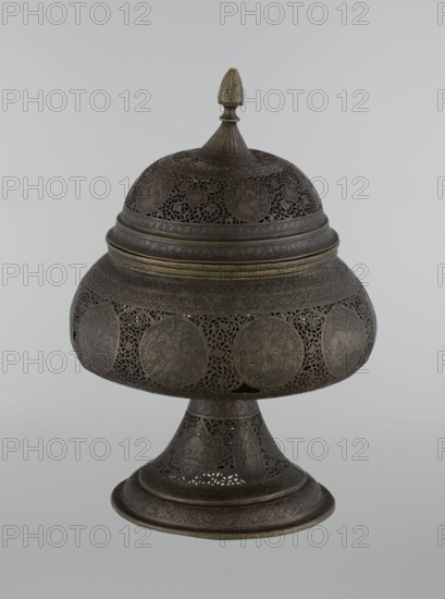 Censer, Persian Iran, Qajar dynasty late 18th–19th century. Brass censer, height 33.0 cm, diameter 22.0 cm. Decorated with figurative scenes and hunting iconography; human figures, animals, demons in cartouches; dense arabesque background; plant motifs; fantastic creatures and beasts dragons, fantastical beasts, riders, acrobatics; Islamic art, Persian decorative style., A domed, lidded metal object on a flared pedestal with elaborate openwork and engraved circular medallions, showing a dark brown patina with areas of darker and lighter brown and subtle greenish-brass highlights, featuring a pointed finial on top, layered bands and filigree patterns around the dome and pedestal, and intricate floral and geometric motifs throughout.