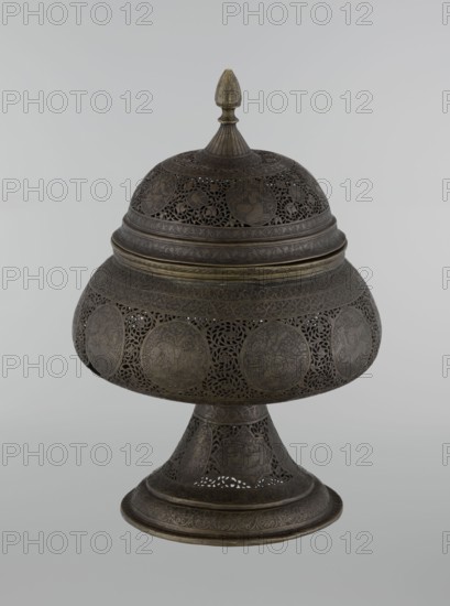 Censer, Persian Qajar dynasty, 18th/19th century, Iran. Brass censer, height 33.0 cm, diameter 22.0 cm. Decoration: figurative scenes including hunting, riders, acrobatics; human figures, animals, demons in cartouches; dense arabesques, plant motifs, fantastic creatures dragons, beasts, fantasy scenes., An ornate metal covered vessel with a domed lid and pointed finial sits against a plain light gray background; the vessel is dark brown with areas of muted gold patina and features intricate openwork filigree and repeated circular medallions around the body, a flared pedestal base with engraved patterns, and a rounded midsection topped by concentric bands and decorative borders, showing a few subtle variations of brown and gold tones.