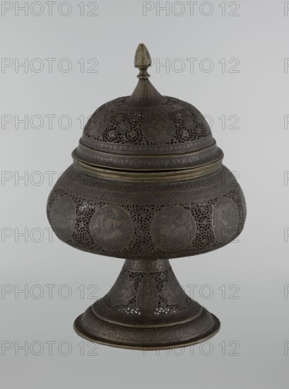 Censer, Persian, Qajar dynasty 1785–1925, 18th–19th century; Iran. Brass censer, height 33.0 cm, diameter 22.0 cm. Decoration: figurative scenes of hunting, human figures, animals, demons in cartouches; dense arabesque background with plant motifs, fantastic creatures dragons, beasts, riders, acrobatics; Islamic art., A decorative metal vessel with a lid and pedestal base sits against a plain light gray background; the vessel is dark brown with areas of muted brass tones and features intricate openwork filigree and rounded medallion panels around its bulbous midsection and dome-shaped lid, and the lid is topped by a small finial that shows a slightly lighter brass color, while the pedestal and rim display repeating engraved patterns and the overall surface shows a subtle aged patina.
