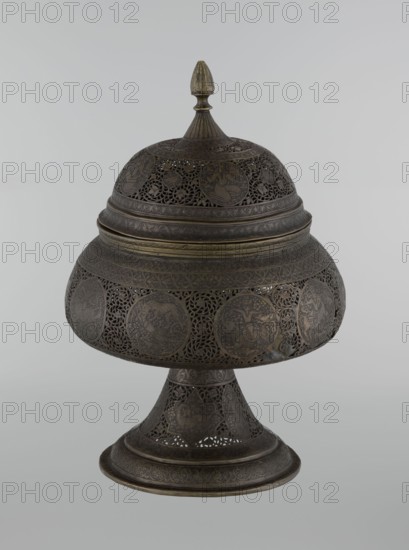 Censer, Persian Iran, Qajar dynasty 1785–1925, 18th/19th century. Bronze censer, height 33.0 cm, diameter 22.0 cm. Decoration: figurative scenes including hunting, human figures, animals, demons in cartouches; dense arabesque background with plant motifs, fantastic creatures dragons, beasts, fantasy scenes, acrobatics, riders., An ornate metal vessel with a domed lid and a pointed finial sits on a flared pedestal base; the surface shows intricate openwork and engraved circular medallions arranged around the dome and body, with repeating floral and geometric patterns and pierced lattice details. The object appears metallic with dark brown and aged brass tones, with subtle variations showing darker patina and lighter golden highlights on raised edges. The overall shape is bulbous at the midsection with a stepped band near the lid, and the pedestal narrows then flares to a wide foot decorated with similar filigree and engraved motifs. The background is a plain light gray.