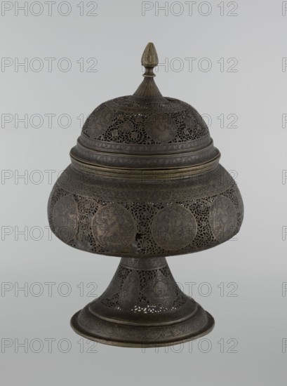 Censer, Persian Iran, Qajar dynasty 1785–1925, 18th/19th century. Metal censer, height 33.0 cm, diameter 22.0 cm. Decoration: figurative scenes including hunting, human figures, animals, demons in cartouches; dense arabesques in the background; fantastic creatures such as dragons and beasts, acrobatics, riders, and plant motifs., A rounded lidded metal vessel on a flared pedestal with intricate openwork and engraved circular medallions, primarily dark bronze with areas of lighter brass or gold-toned highlights, showing a domed top with a pointed finial, a wide midsection with pierced floral and geometric patterns, and a tapered base decorated with similar filigree motifs.