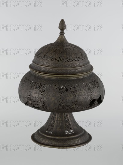 Censer, Persian, Qajar dynasty 1785–1925, 18th–19th century, Iran. Brass censer, height 33.0 cm, diameter 22.0 cm. Decoration: figurative scenes including hunting, human figures, riders, acrobatics; animals, dragons and fantastical beasts, demons in cartouches; dense arabesques and plant motifs; fantasy scenes., An ornate metal container with a domed lid and finial sits on a flared pedestal; the object displays intricate openwork and engraved patterns across its rounded body, lid, and base, with decorative panels featuring stylized figures and ornamental motifs visible through the filigree. The surface shows dark bronze tones with subtle variations of brown and muted gold highlights, and the background is a smooth pale gray.