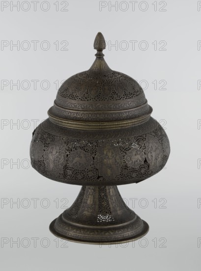 Censer, Persian, Qajar dynasty late 18th–19th century, Iran. Brass censer with figurative scenes of hunting, riders, acrobatics, dragons and fantastic beasts, human figures, animals and demons in cartouches, dense arabesque background and plant motifs; height 33.0 cm, diameter 22.0 cm., An ornate metal lidded vessel with a rounded body and pedestal base, displaying intricate openwork and pierced patterns; the lid has a pointed finial and layered decorative bands, and the body shows a series of framed figural and animal motifs within delicate filigree; the pedestal flares outward and repeats the pierced and engraved decoration; visible colors include dark brown, bronze, and hints of muted gold against a pale gray background.