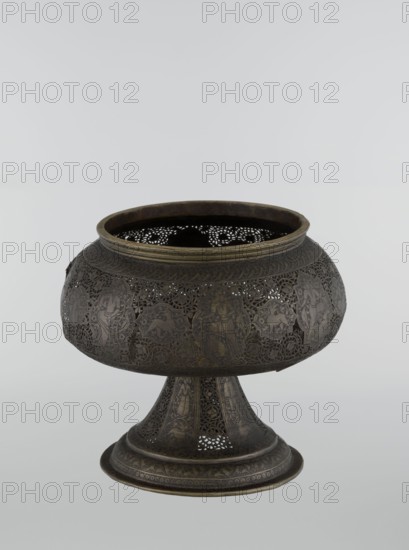 Censer, Persian Iran, Qajar dynasty late 18th–19th century. Bronze censer, height 33.0 cm, diameter 22.0 cm. Decoration: figurative scenes including hunting, riders, acrobatics; human figures, animals, demons in cartouches; dense arabesque background; plant motifs; fantastic creatures such as dragons and beasts; fantasy scenes., A low, rounded metal vessel on a flared pedestal with an open top, featuring intricate pierced and engraved patterns across its bulbous body and base; the surface shows a dark brown and black patina with areas of muted gold or brass tones, and the interior appears dark.