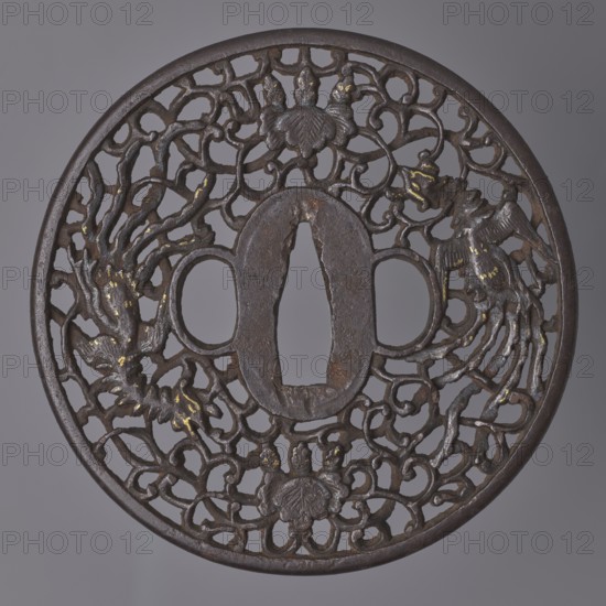Crossguard tsuba, maker, images of birds; Japanese sword crossguard tsuba, namban style; materials: paulownia bot., gold nunome inlay; iconography: hoo phoenix / hôô phoenix and paulownia kiri flowers woven into a karakusa twig; openwork decoration; function: katana crossguard; form: tsuba round maru-gata with rounded edge maru mimi, seppa dai oval and corresponding ryôhitsu ana oval; Edo period, 18th century 1603–1868, Japan; diameter 9.3 cm, thickness 0.5 cm., A round dark metal object with an openwork lattice of curving organic shapes and decorative motifs, showing a central oblong opening and two smaller rounded openings on either side; the metal surface is predominantly dark brown with areas of lighter brown and rust, and small accents of dull gold scattered across raised details; the design includes a few stylized floral or shell-like elements and two bird-like forms integrated into the openwork, all set against a neutral gray background.