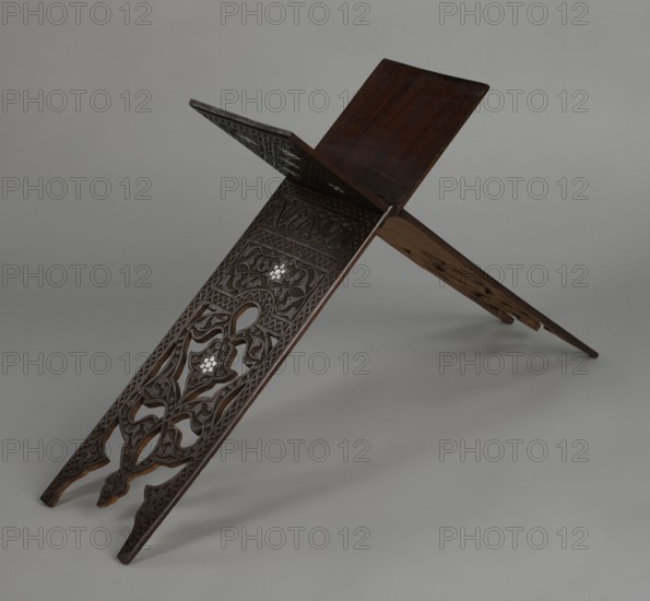 Quran lectern rahla, Ottoman Turkey, 19th century. Wooden piece of furniture decorated with arabesques, geometric ornament, rosettes, lotus tulip motif, lotuses, tulips. Function: Quran lectern. Unfolded: height 50.6 cm, width 91.5 cm, depth 21.3 cm, weight 1.7 kg. Folded: height 80.5 cm, width 21.3 cm, weight 1.7 kg., A carved wooden folding stand with two interlocking panels arranged in an X shape, showing a dark brown finish with lighter brown edges and worn areas; the front panel features ornate openwork carving with flowing geometric and vegetal patterns and inlaid lighter pieces that appear pale or off-white; the top surfaces of the panels are smoother and darker brown, while the background is a smooth neutral gray.