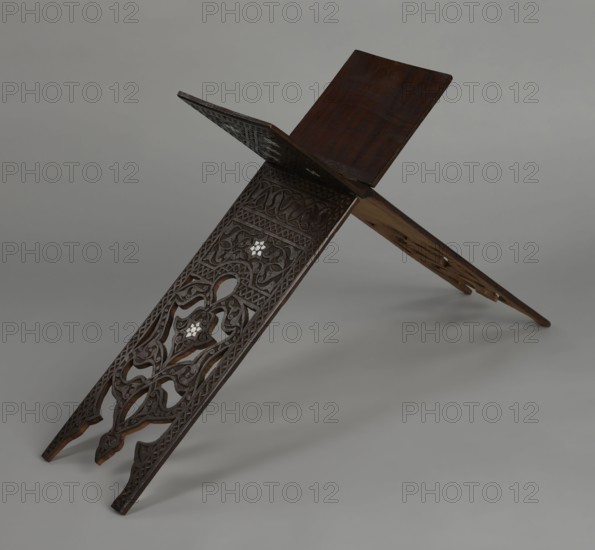 Quran lectern rahla, Ottoman Turkish, 19th century. Wooden piece of furniture used for holding the Quran, decorated with arabesques, geometric ornament, rosettes, lotus tulip motifs, lotuses, and tulips. Decorated with rosettes, geometric ornament, arabesque, lotus tulip motif. Unfolded: height 50.6 cm, width 91.5 cm, depth 21.3 cm, weight 1.7 kg. Folded: height 80.5 cm, width 21.3 cm, weight 1.7 kg., A carved wooden book stand formed by two interlocking panels arranged in an X shape, showing intricate openwork and relief carvings with floral and geometric motifs; the wood is dark brown with areas of lighter brown on the underside and edges, and there are small inlaid pieces that appear white; the background is a smooth gray surface and gray backdrop.